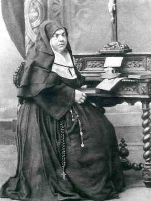 Święta Maria Soledad Torres Acosta siedząca w stroju zakonnym przy stoliku na czarno-białej fotografii.